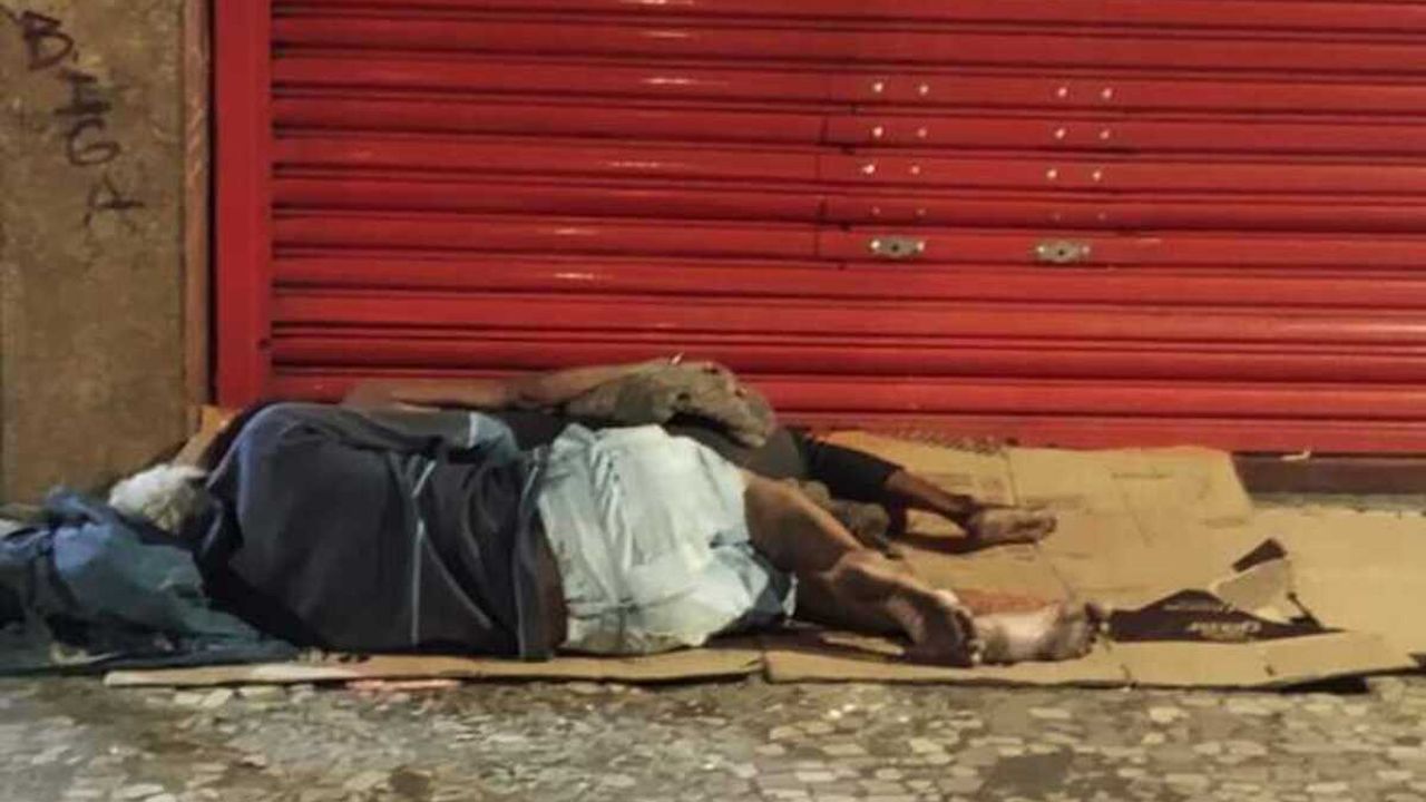 Pessoas dormindo na rua em frente a uma estrutura comercial fechada, com sinalização grafitada.