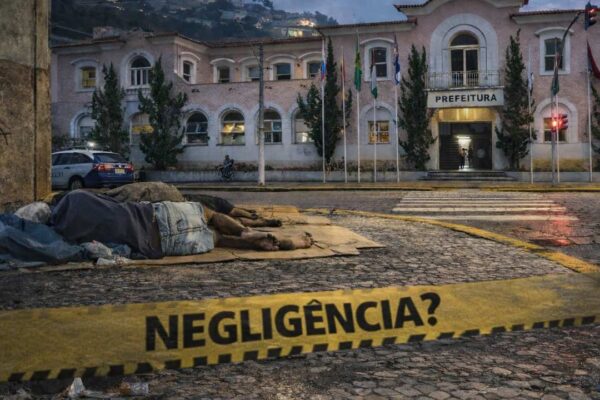 Pessoas em situação de rua dormem em frente à prefeitura. Faixa de alerta com a palavra 'Negligência'.