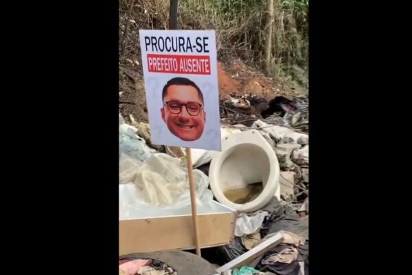 Placa com rosto de político e frase 'Procura-se Prefeito Ausente' em meio a lixo.