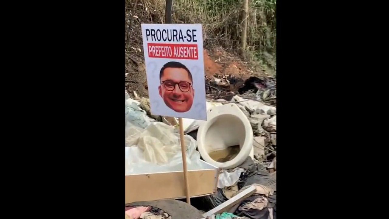 Placa com rosto de político e frase 'Procura-se Prefeito Ausente' em meio a lixo.