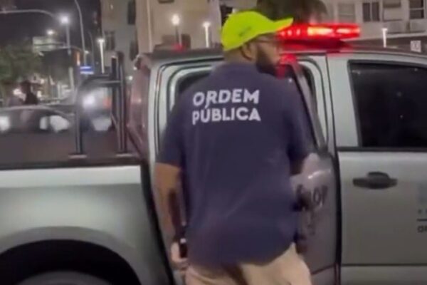 Policial com uniforme 'Ordem Pública' em frente a viatura.