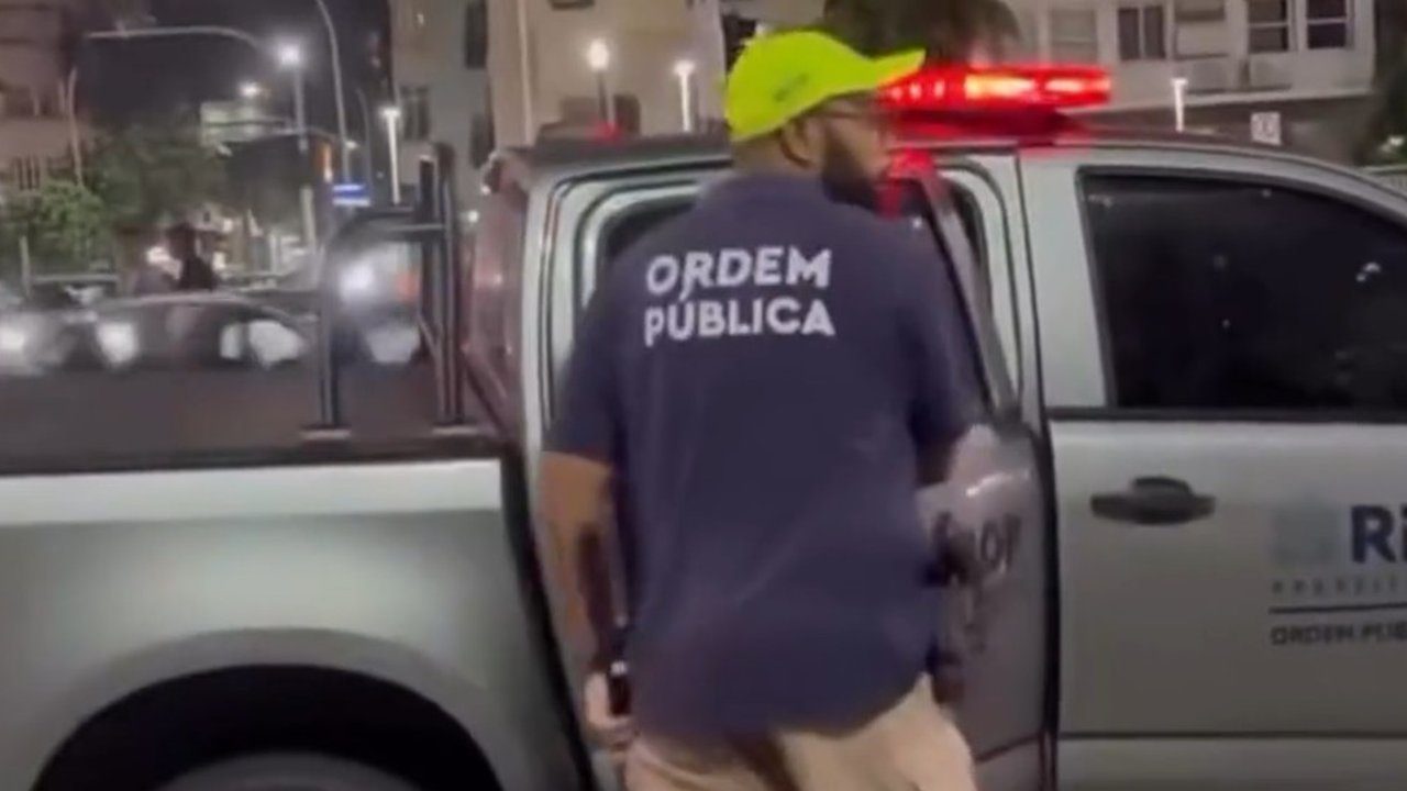 Policial com uniforme 'Ordem Pública' em frente a viatura.