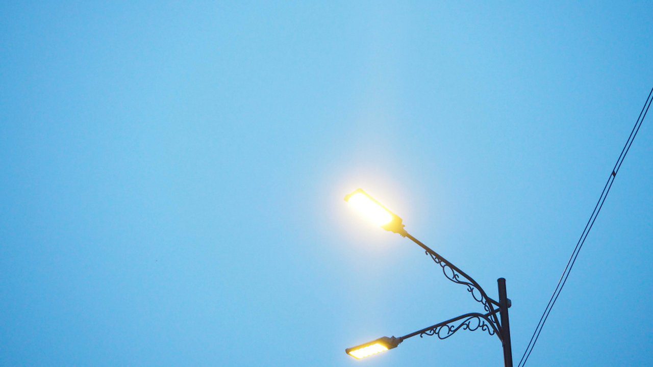 Postes de iluminação pública com luzes acesas contra céu azul.