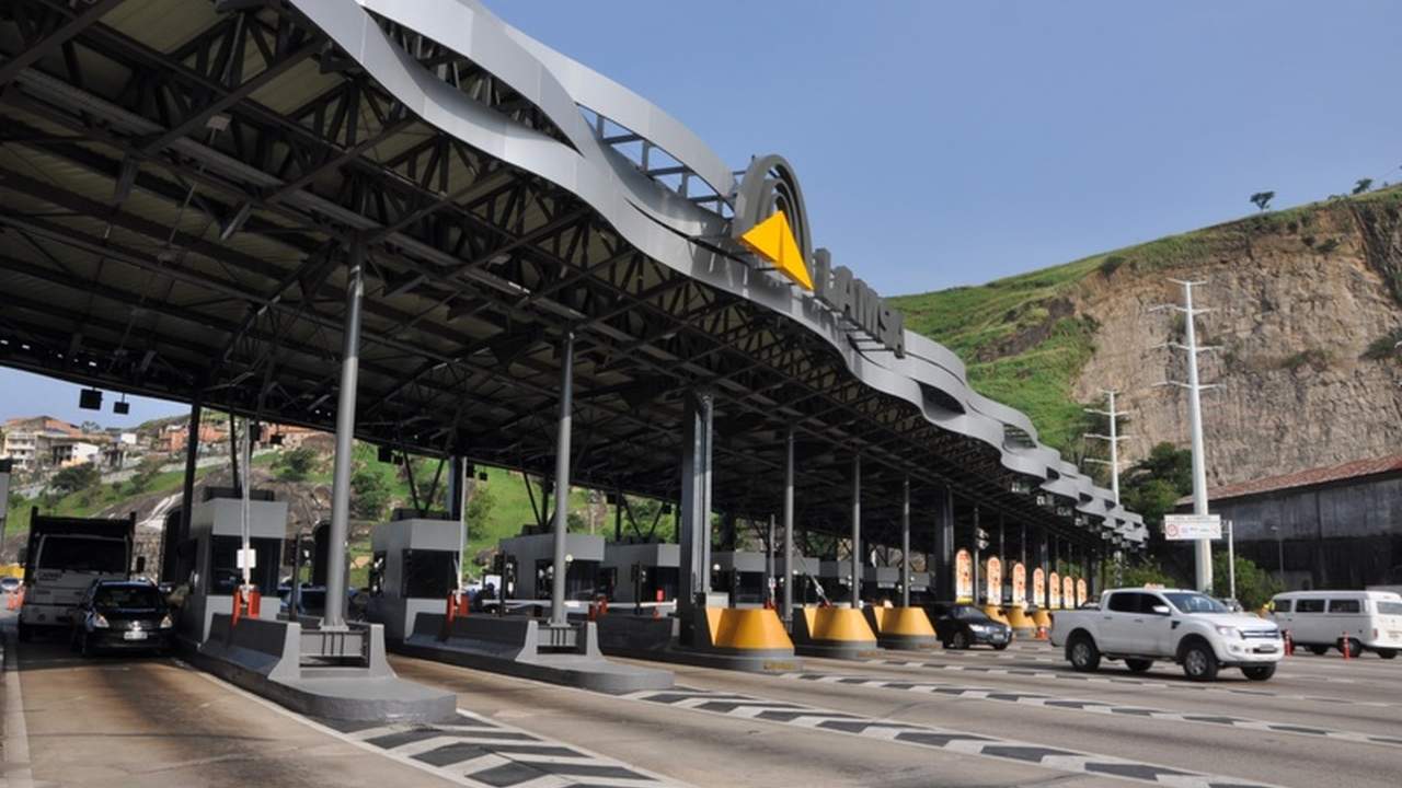 Praça de pedágio em rodovia, com veículos passando e estrutura de cobertura.