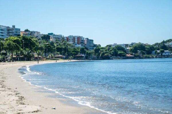 Praia com areia clara e mar azul, com edifícios ao fundo.