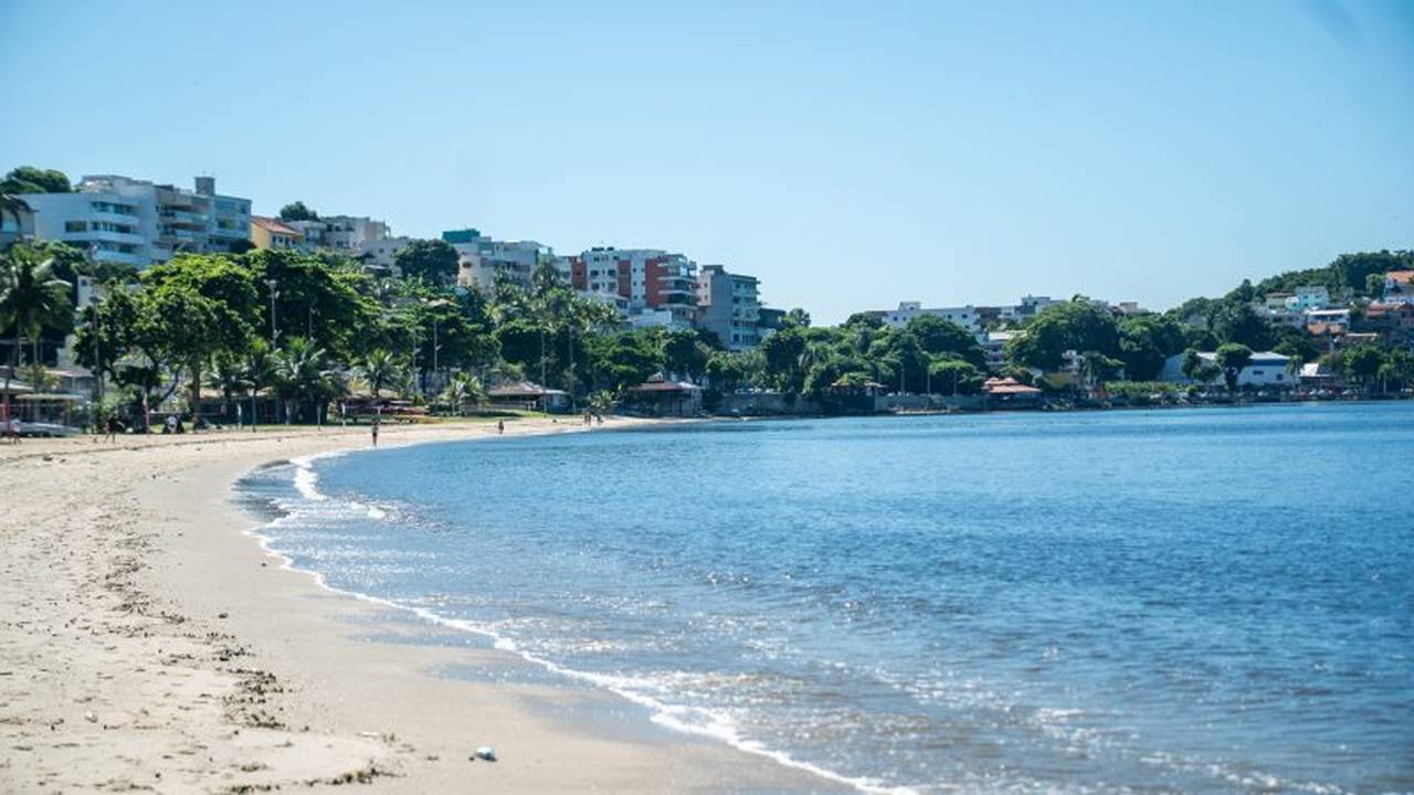 Praia com areia clara e mar azul, com edifícios ao fundo.