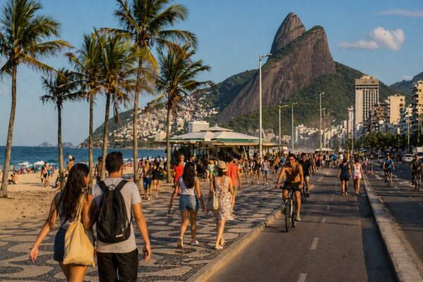 Praia de Ipanema no Rio de Janeiro, com pessoas caminhando e montanhas ao fundo.