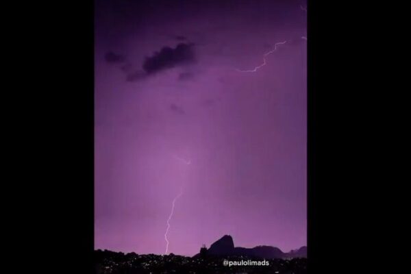 Raios iluminam o céu roxo sobre o Rio de Janeiro em noite de tempestade, com silhuetas urbanas e do mar