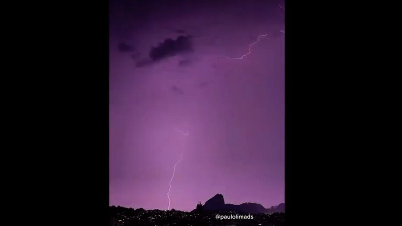 Raios iluminam o céu roxo sobre o Rio de Janeiro em noite de tempestade, com silhuetas urbanas e do mar