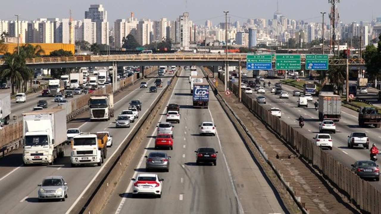 Rodovia com tráfego intenso e prédios ao fundo.