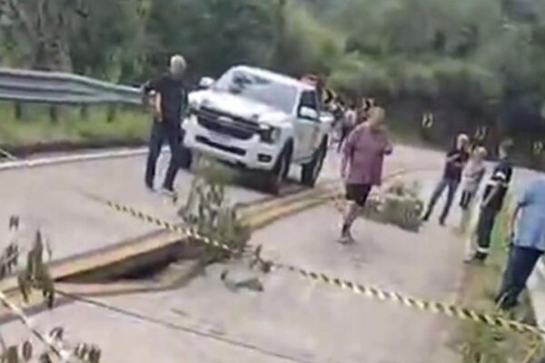 Rodovia interditada após parte da pista ceder, com carro parado e pessoas ao redor.