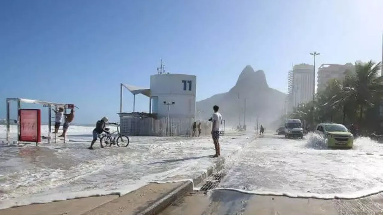 Rua alagada no Rio de Janeiro devido à ressaca marítima, com carros e pessoas afetadas.
