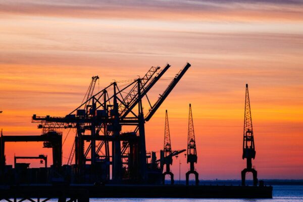 Silhouette de guindastes portuários contra o céu alaranjado ao pôr do sol.