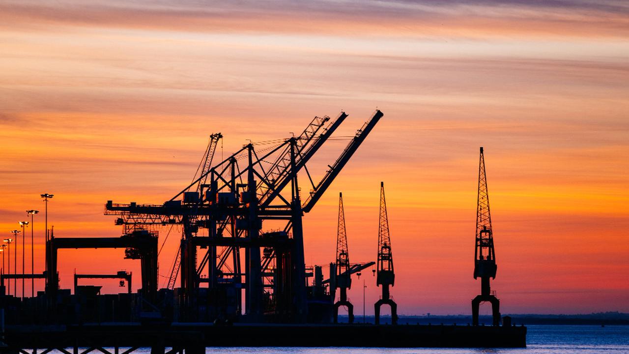 Silhouette de guindastes portuários contra o céu alaranjado ao pôr do sol.