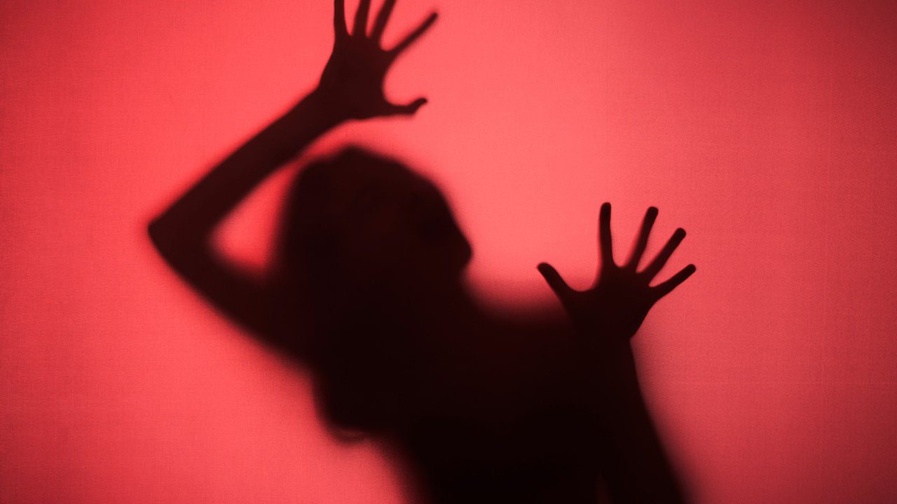 Sombra de pessoa com mãos estendidas em fundo vermelho, simbolizando violência.
