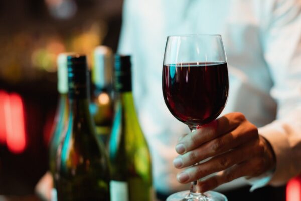 Taça de vinho tinto sendo oferecida, com garrafas de vinho ao fundo e um garçom.