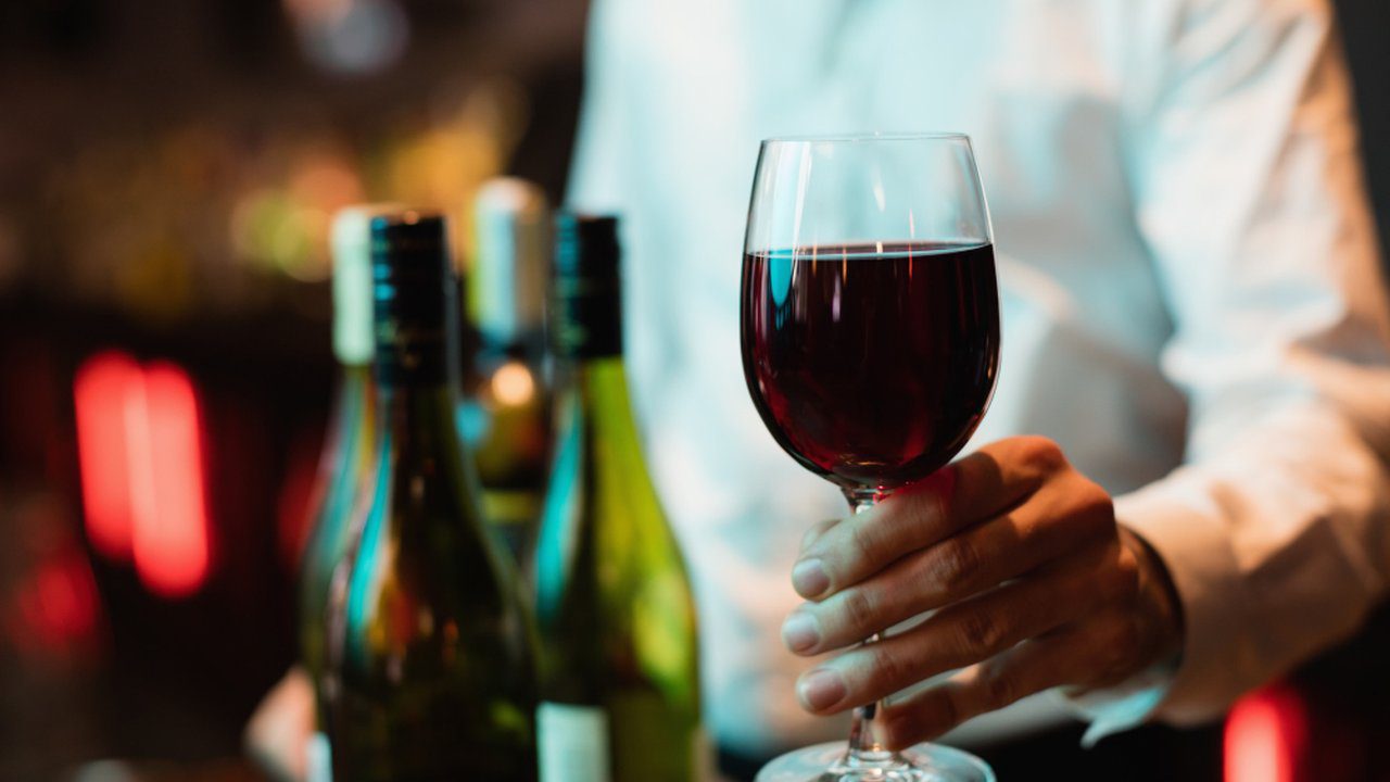 Taça de vinho tinto sendo oferecida, com garrafas de vinho ao fundo e um garçom.