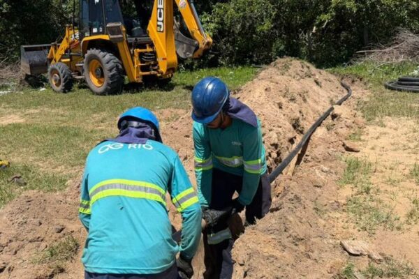 Trabalhadores instalando tubulação em obra, com máquina JCB ao fundo.