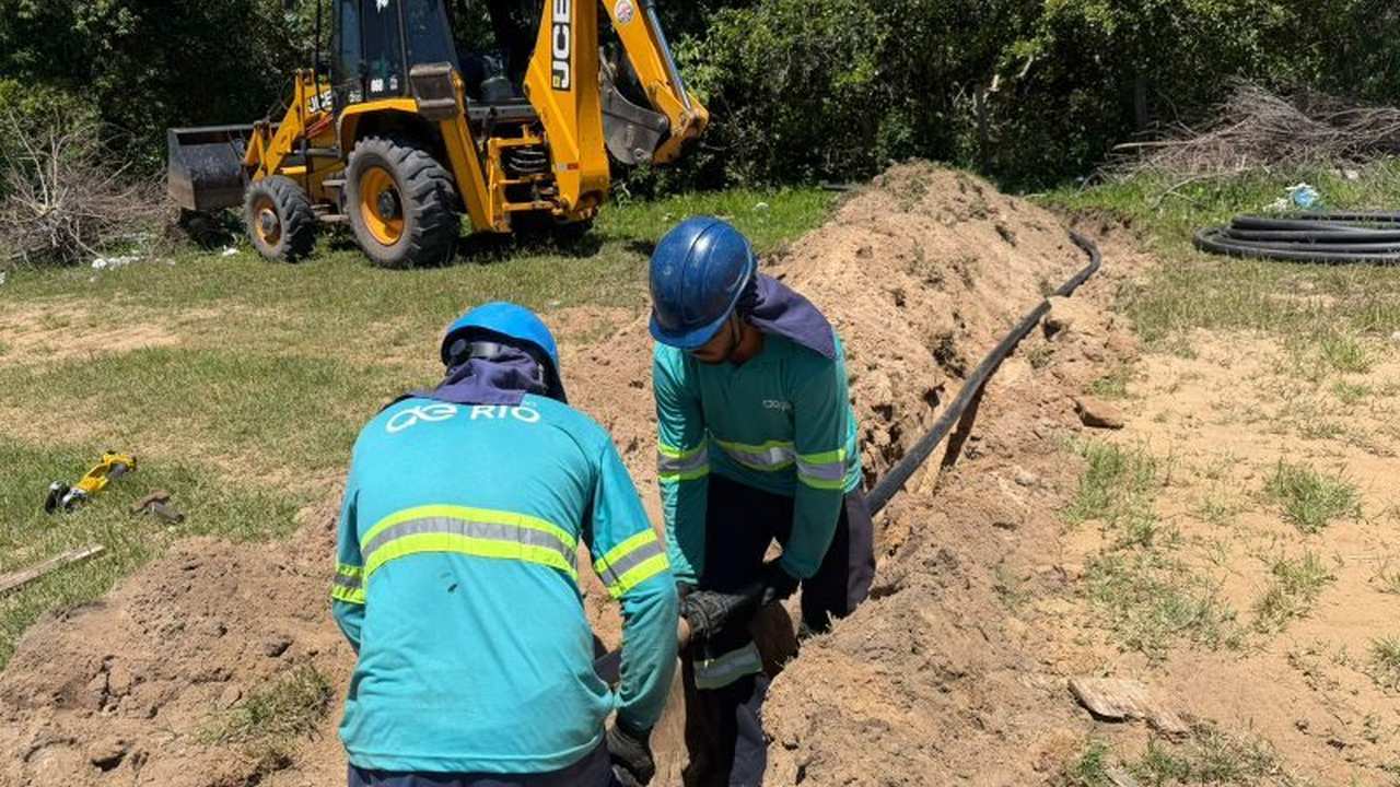 Trabalhadores instalando tubulação em obra, com máquina JCB ao fundo.