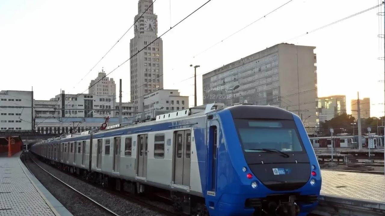 Trem azul e branco em estação no Rio de Janeiro, com prédios ao fundo.
