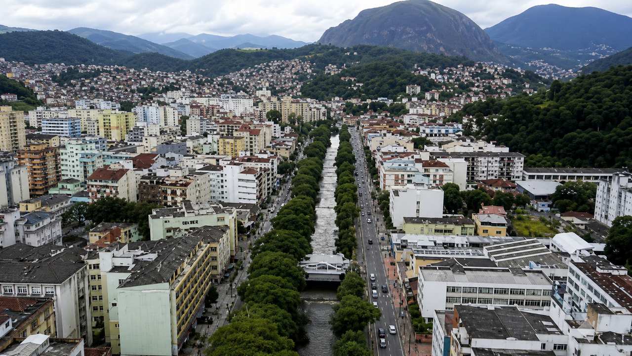 Vista aérea de Nova Friburgo, com prédios, rio e montanhas ao fundo.