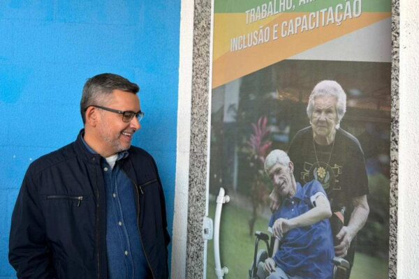 Zé Alexandre sorrindo ao lado de banner sobre inclusão e capacitação, com imagem de pessoas com deficiência.