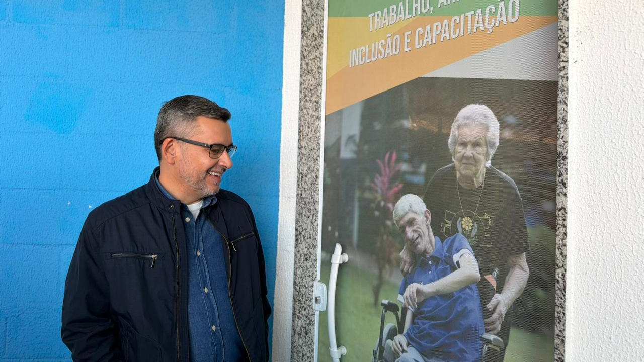 Zé Alexandre sorrindo ao lado de banner sobre inclusão e capacitação, com imagem de pessoas com deficiência.