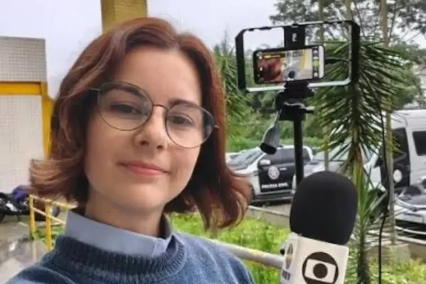Juliana Guzzo segura um microfone durante cobertura externa, com celular montado em tripé ao fundo.