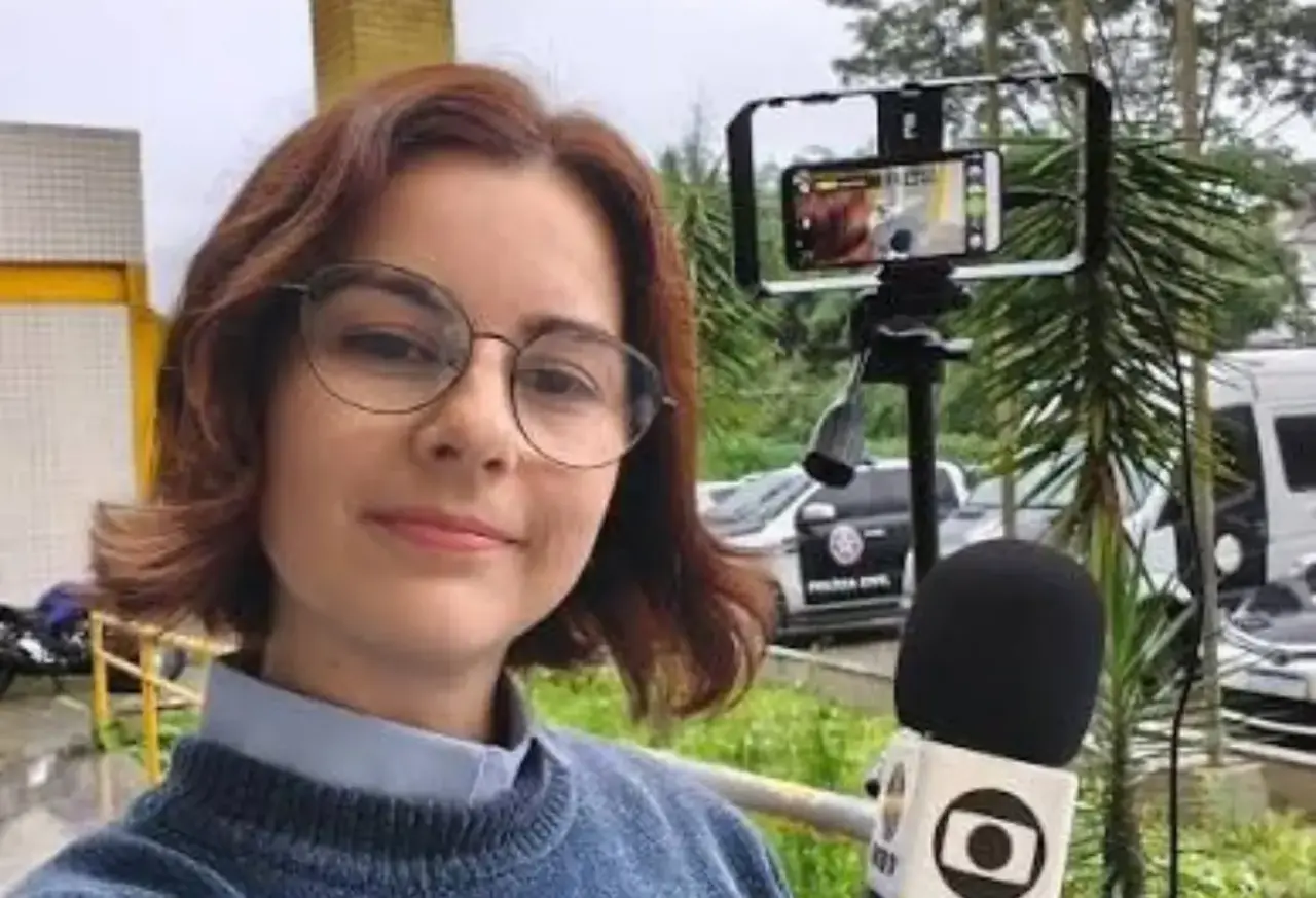 Juliana Guzzo segura um microfone durante cobertura externa, com celular montado em tripé ao fundo.