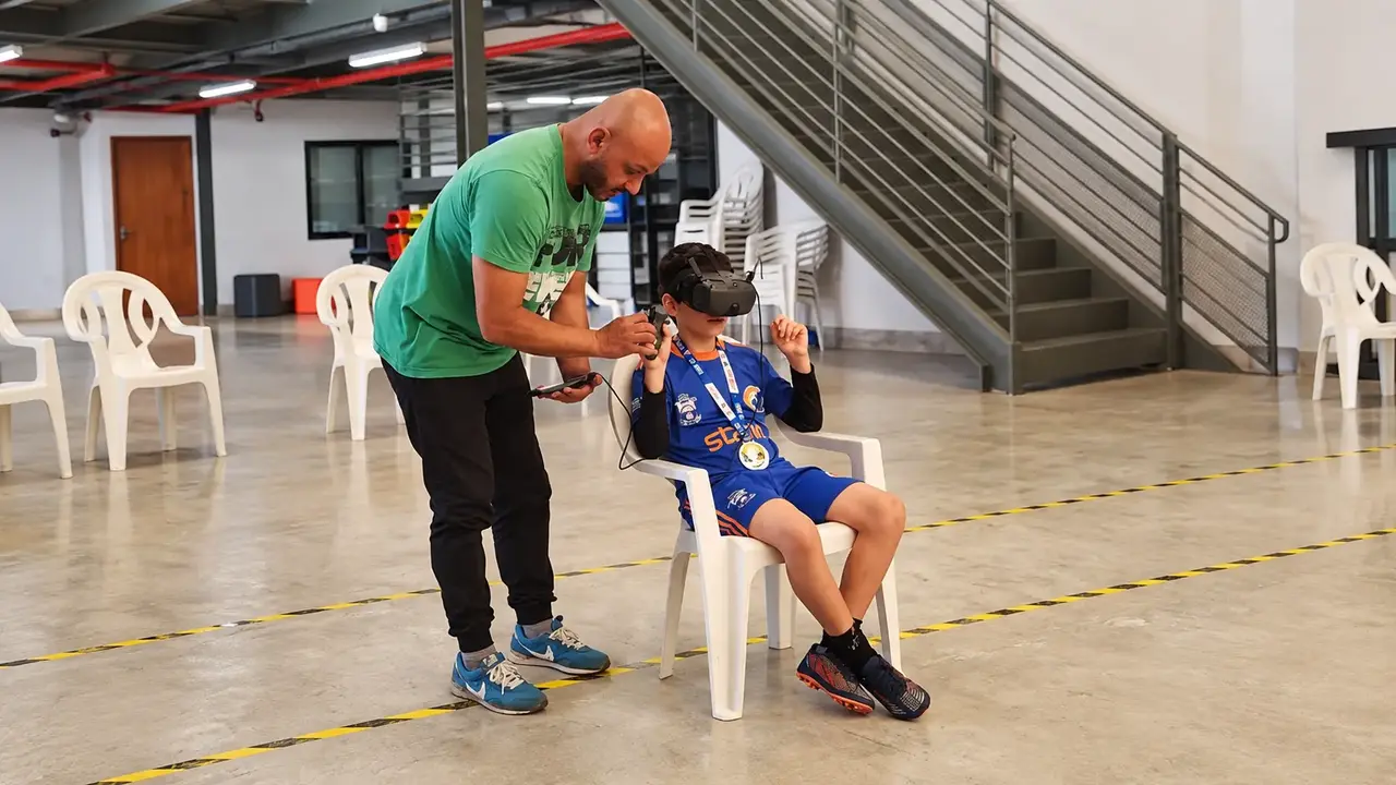 Adulto auxilia criança sentada a experimentar equipamento com visor durante atividade no Sesc Nova Friburgo