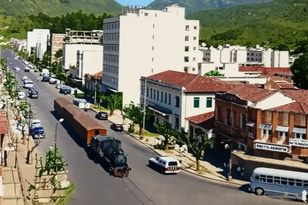 Trem com vagões de carga percorre uma avenida de Nova Friburgo entre carros e edificações.
