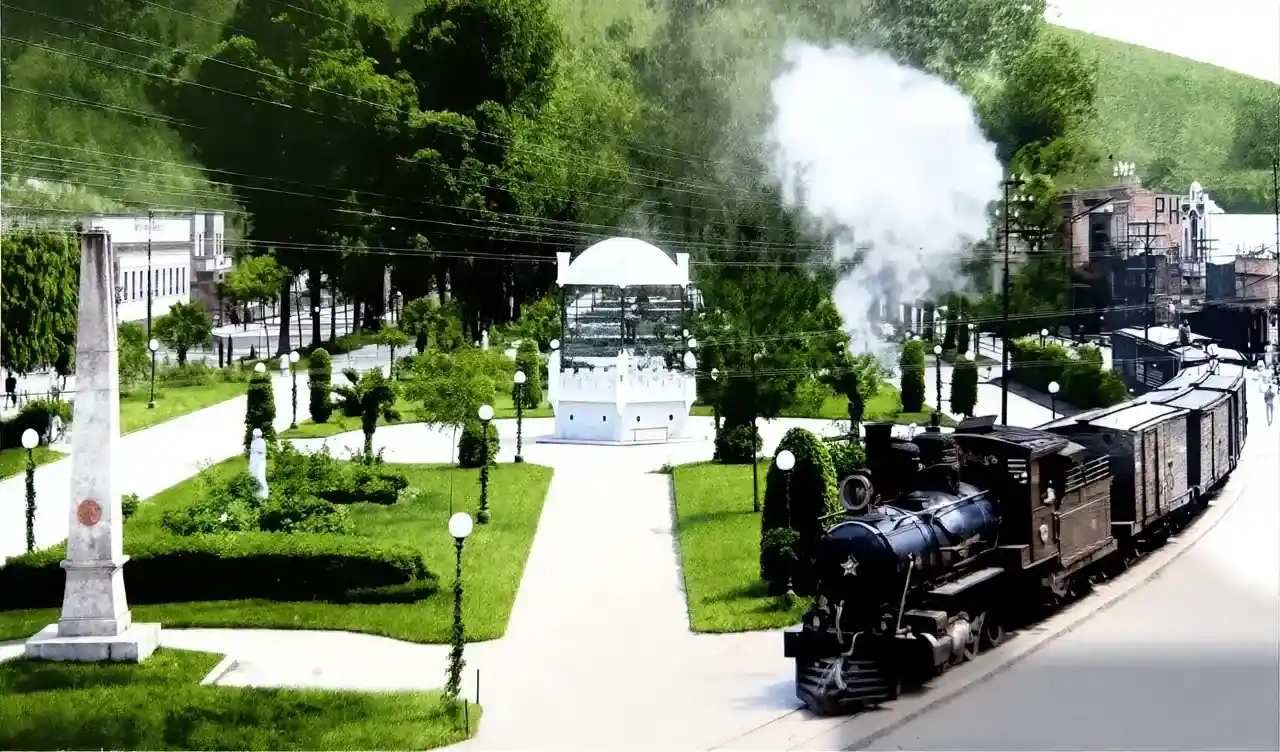 Trem a vapor passa ao lado de uma praça com jardins, árvores, obelisco e coreto em Nova Friburgo.