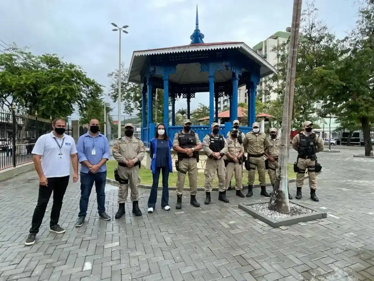 Grupo de pessoas, incluindo agentes uniformizados, posa diante de um coreto azul em uma praça.