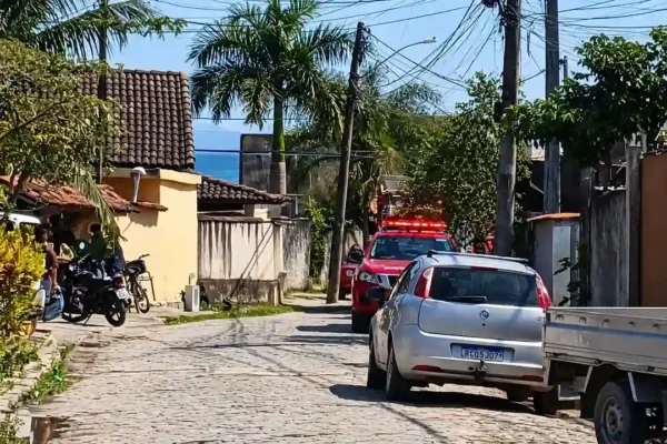 Rua residencial com viatura vermelha de resgate, veículos estacionados, moradores e postes com fiação aérea.