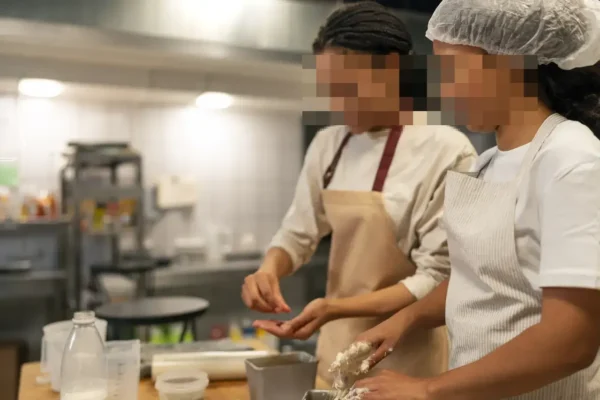 Duas mulheres de avental preparam alimentos em uma cozinha, com os rostos pixelizados.