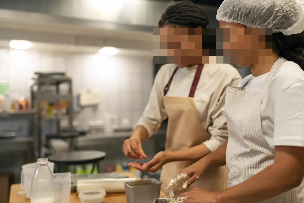 Duas mulheres de avental preparam alimentos em uma cozinha, com os rostos pixelizados.