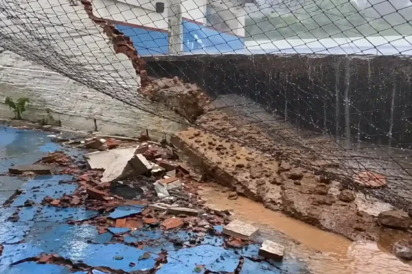 Muro rompido e tela de proteção inclinada sobre escombros e água em escola no Caleme.