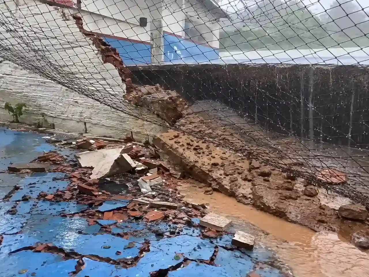 Muro rompido e tela de proteção inclinada sobre escombros e água em escola no Caleme.