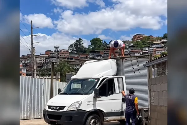 Caminhão branco junto a uma barra limitadora de altura, com um homem sobre a cabine e outro ao lado da porta aberta.