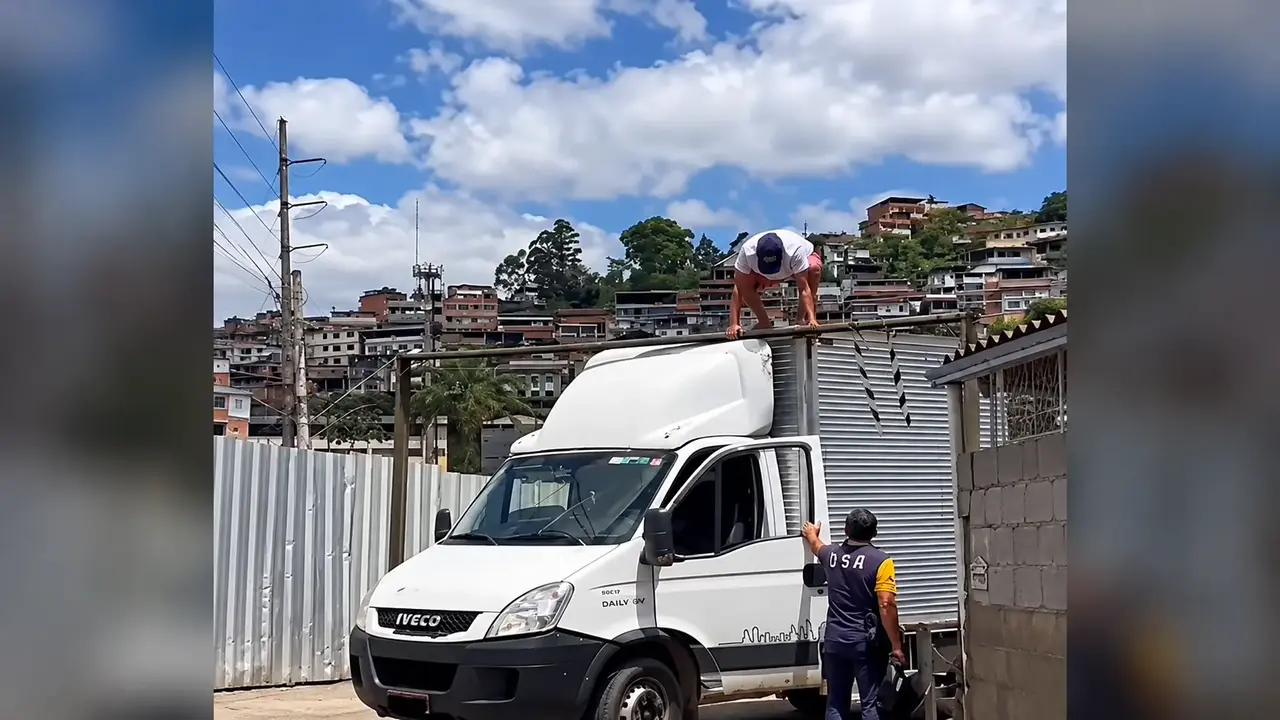Caminhão branco junto a uma barra limitadora de altura, com um homem sobre a cabine e outro ao lado da porta aberta.