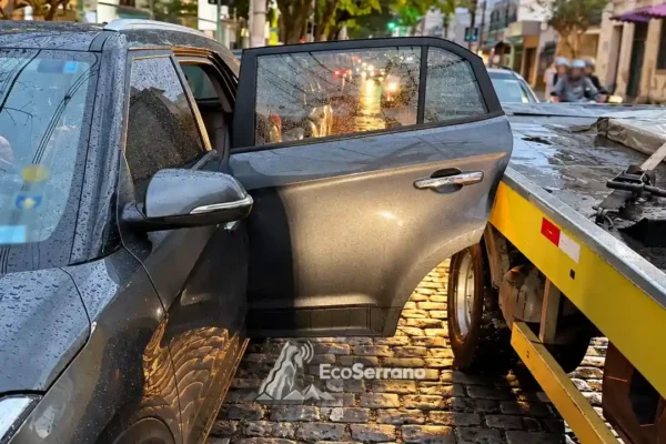 Porta lateral de carro cinza junto à plataforma de um caminhão-guincho, em rua com pavimento molhado.