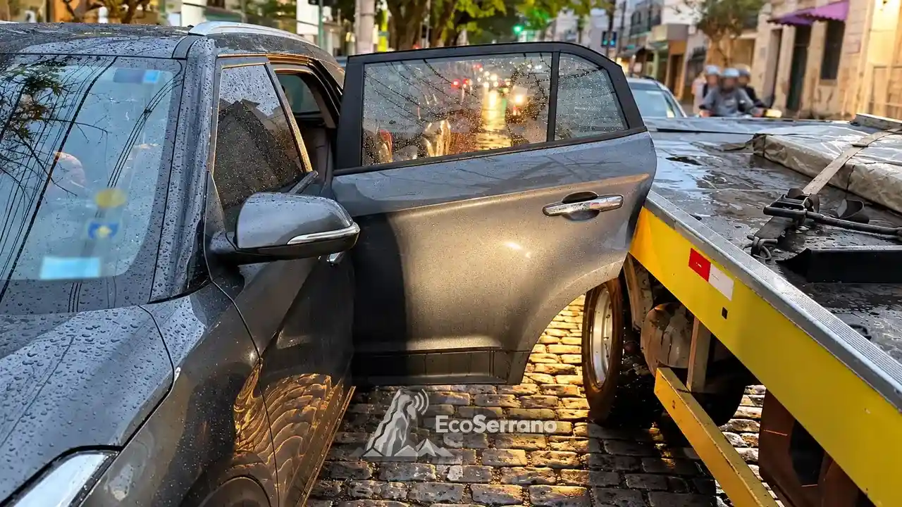Porta lateral de carro cinza junto à plataforma de um caminhão-guincho, em rua com pavimento molhado.