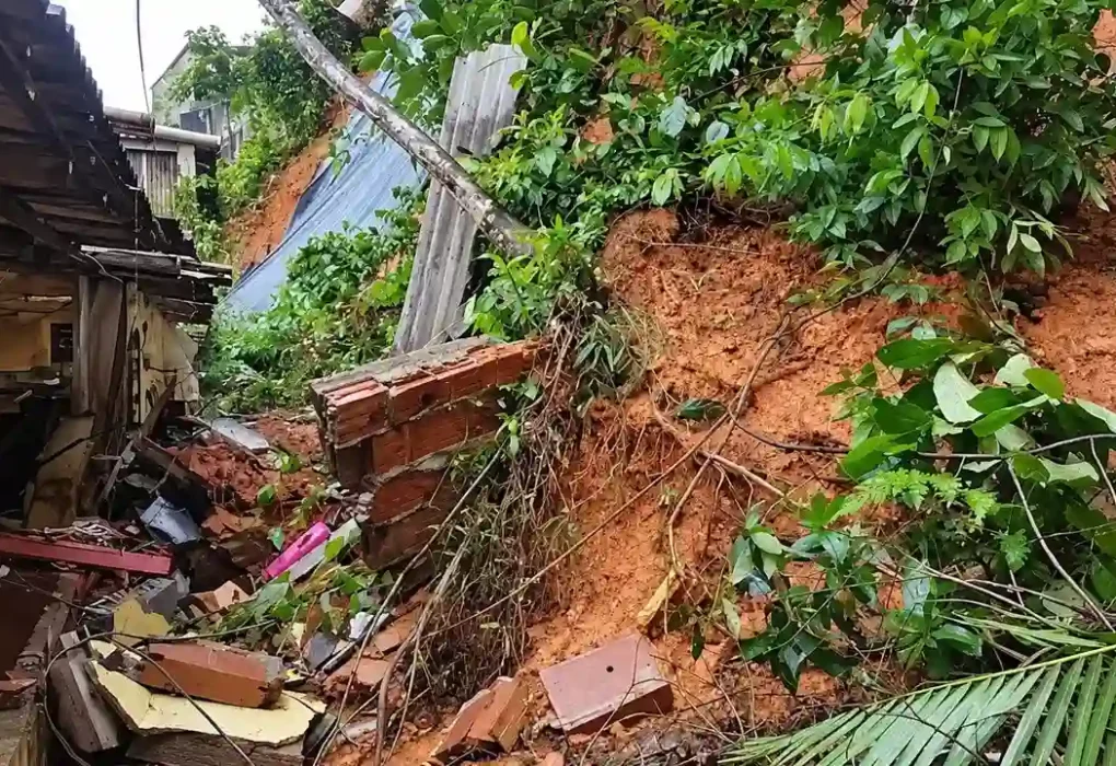 Encosta com terra exposta, vegetação caída e escombros ao lado de uma residência.