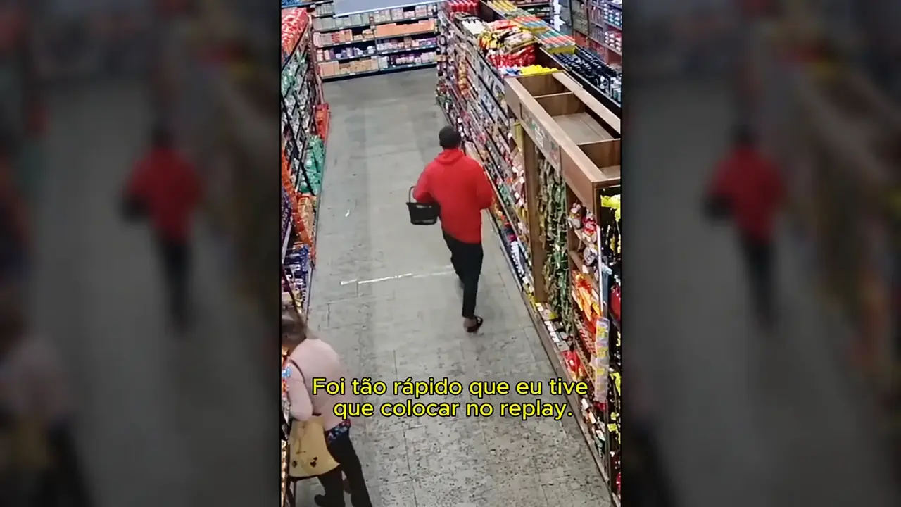 Imagem de câmera de segurança mostra pessoa de roupa vermelha carregando uma cesta em corredor de supermercado.