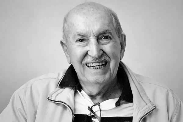 Carlos Lyra sorrindo em retrato em preto e branco, usando jaqueta clara e óculos pendurados na camisa.
