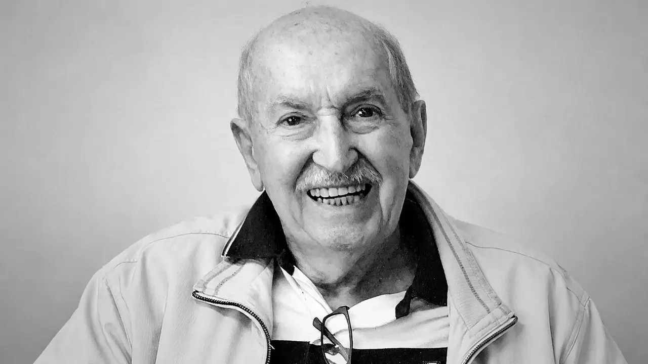 Carlos Lyra sorrindo em retrato em preto e branco, usando jaqueta clara e óculos pendurados na camisa.