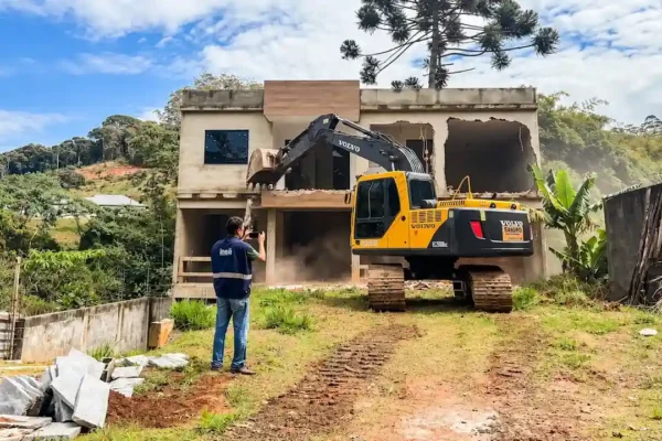 Escavadeira amarela junto aos escombros de uma casa, em terreno próximo à vegetação e a um córrego.