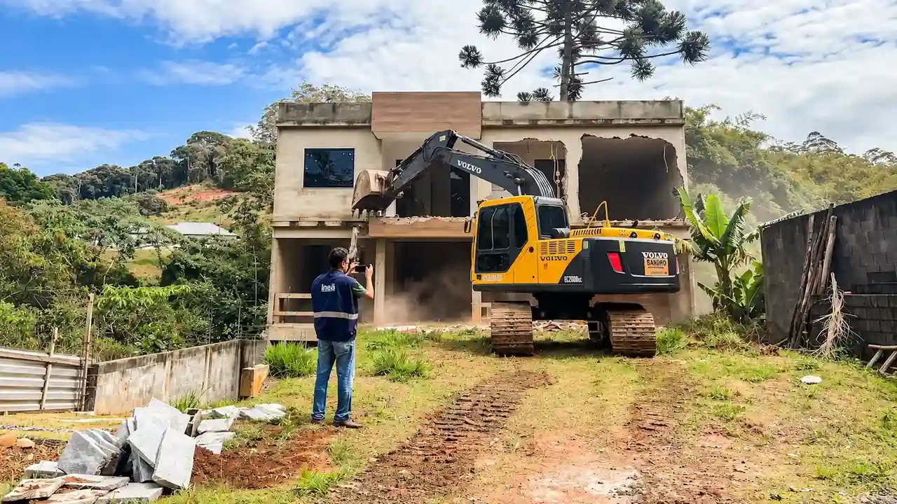 Escavadeira amarela junto aos escombros de uma casa, em terreno próximo à vegetação e a um córrego.