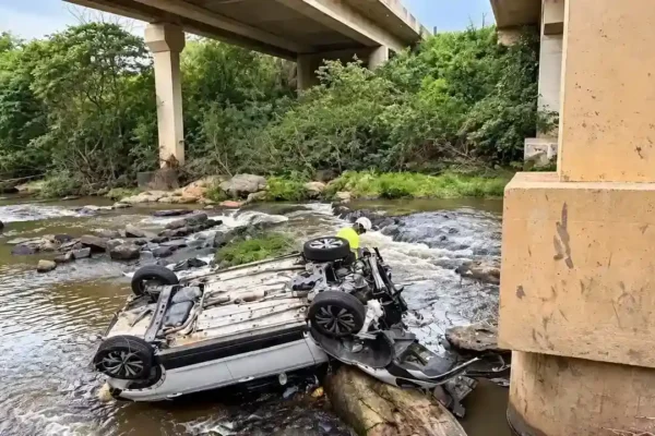 Carro capotado em um curso d’água sob uma ponte, com uma pessoa próxima ao veículo.