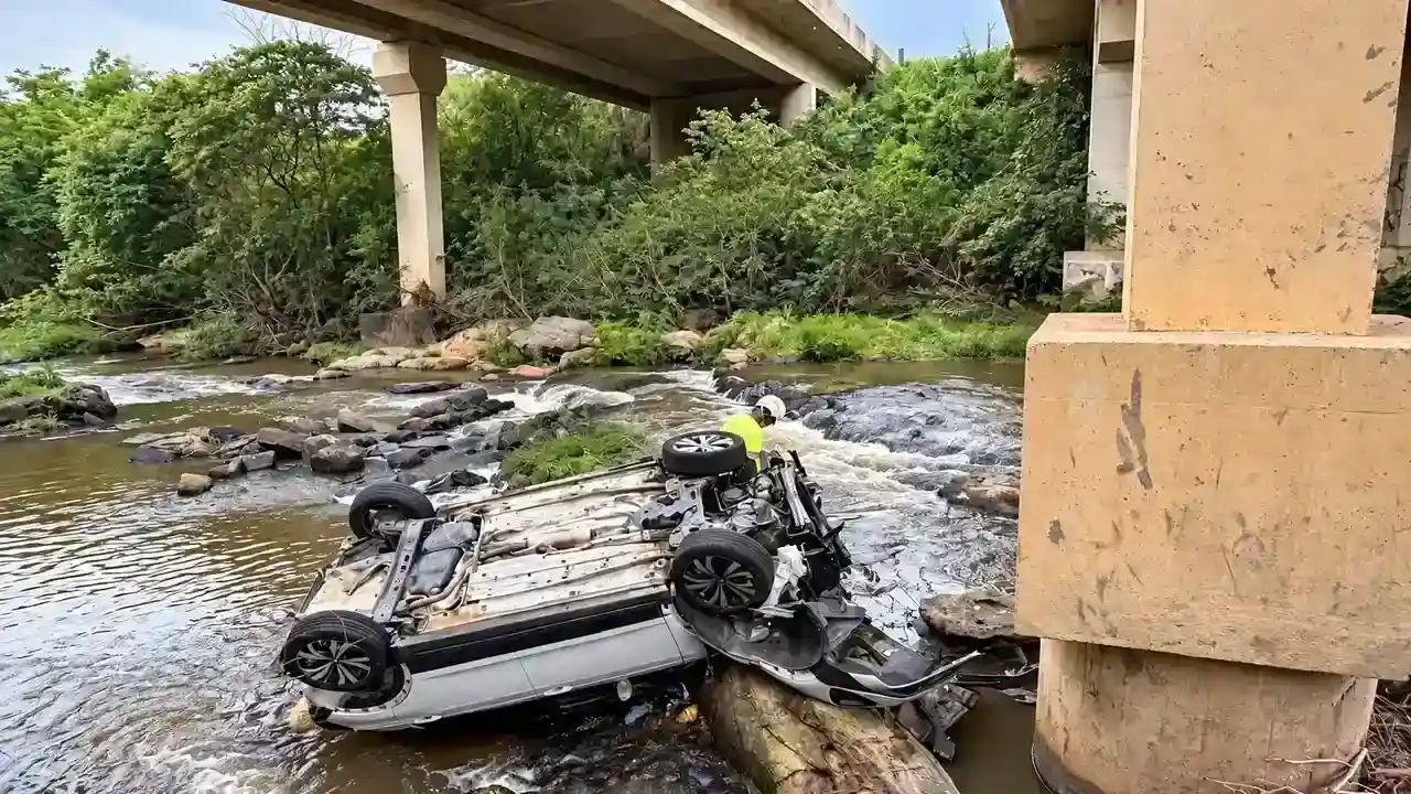 Carro capotado em um curso d’água sob uma ponte, com uma pessoa próxima ao veículo.